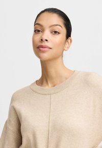 Beige Strickpullover mit rundem Halsausschnitt, gerippten Bündchen und einer Nahtdetails auf der Vorderseite. Weicher Stoff und bequeme Passform.