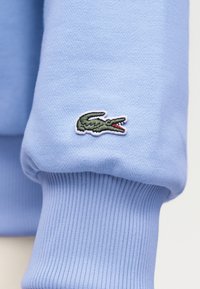 Manche tricotée bleu clair avec poignet côtelé et petit logo crocodile vert brodé près du poignet.