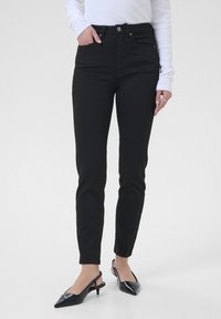 Jean taille haute noir avec une coupe slim. Tissu en m�élange de coton, poches avant et fermeture à bouton. Associé à des escarpins noirs à talon slingback.