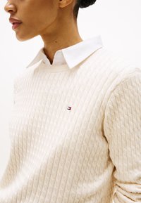 Crème kabelgebreide trui met ronde hals, met daaronder een witte blouse. De trui heeft een klein logo-accent op de borst.