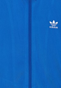 Modrá bunda na zip vyrobená z lehkého materiálu, s bílým vyšívaným logem Adidas na horní levé straně.