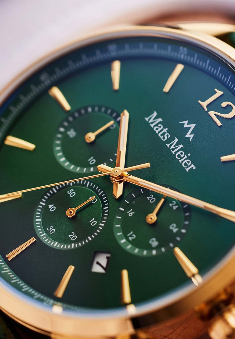 Goldfarbene Uhr mit grünem Zifferblatt, ausgestattet mit drei Teilzifferblättern, goldenen Stundenmarkierungen und einem Datumsdisplay. Glatte, polierte Oberfläche.
