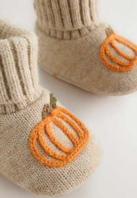 Chaussons pour bébé tricotés beige avec des bords côtelés et des motifs de citrouilles orange sur les orteils, posés sur une surface blanche.
