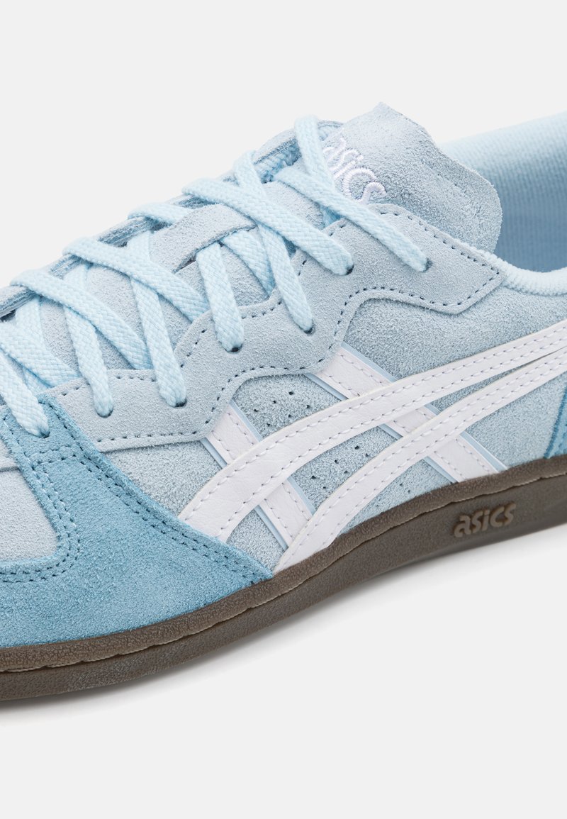 Zapato atlético de ante azul claro con detalles en cuero blanco, cordones planos y suela de goma texturizada con marcas visibles en el lateral.