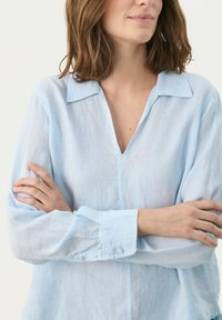 Camicia in lino azzurro chiaro con scollo a V, maniche lunghe e polsini con bottone. Presenta un colletto classico e una vestibilità rilassata. Texture morbida.