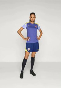 Athlète féminine se tenant avec confiance, portant un maillot de soccer bleu et jaune de l'Atlético de Madrid, avec des chaussettes noires et des crampons, sur un fond uni.