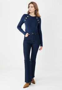 Granatowy sweter z dzianiny o żebrowej fakturze, z złotymi guzikami na ramionach, w połączeniu z high-waisted ciemnoniebieskimi dżinsami w stylu dzwonów i brązowymi botkami na obcasie.