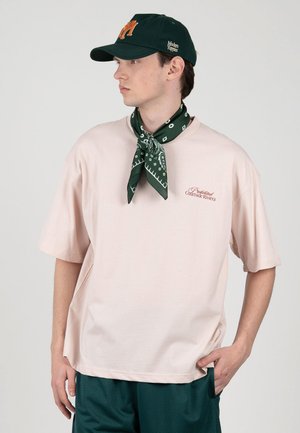 Young man wearing light pink oversized T-shirt, dark green baseball cap, matching neck scarf, and dark green shorts, looking to the side.