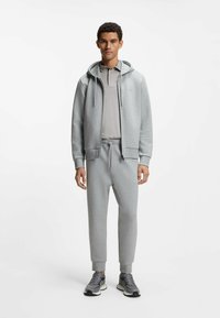 Lysegrå hættetrøje med lynlås og snore, kombineret med en matchende langærmet skjorte og tætsiddende joggingbukser. Grå sneakers fuldender looket.