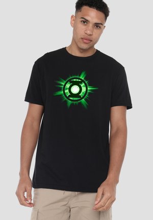 Young man wearing a black t-shirt with a glowing green Green Lantern emblem, paired with beige pants, standing against a plain background.