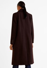 Dlouhý, tmavě hnědý kabát vyrobený z měkké tkaniny, se rovný střihem, výstřihem do V a knoflíkovými manžetami. Minimální detaily, jednoduchý design.