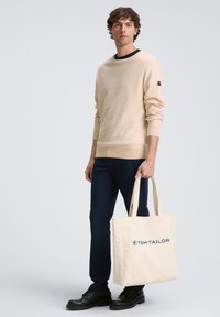 Bēša džemperis ar tumšu apkakli, tumši zili svārki un melni apavi; rokā rokassoma krēmkrāsā ar "TOM TAILOR" zilu logotipu.