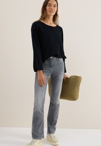 Zwarte blouse met lange mouwen en een ronde hals, gecombineerd met lichtgrijze recht-toe-recht-aan jeans. Houdt een klein, olijfgroen kussen vast.