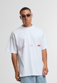 Bílý bavlněný tričko s krátkým rukávem, které na přední straně a detailu bočního švu obsahuje červený grafický text "Another" a "ACL".