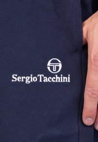 Tissu bleu marine de Sergio Tacchini avec logo brodé blanc. Main positionnée sur la couture latérale près du logo, mettant en valeur la texture du matériau et les détails de conception.