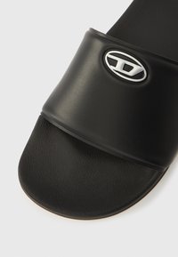 Sort slidessandal med en glat, mat rem med en sølv oval logo. Fodbedet har en struktureret overflade og afrundede kanter.