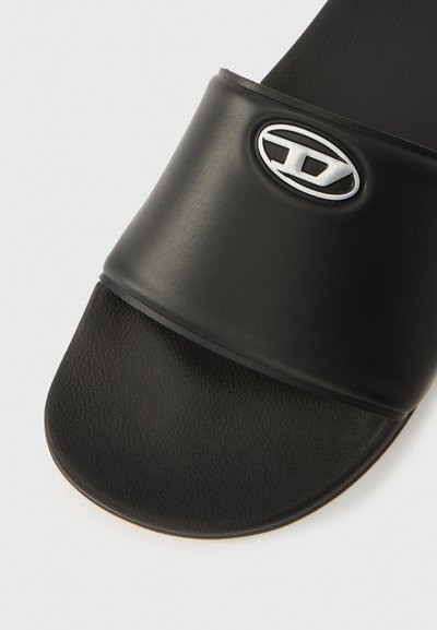 Sandale à glissière noire avec une bride lisse et mate ornée d'un logo ovale argenté. La semelle intérieure présente une finition texturée et des bords arrondis.