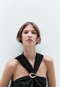 Černý halter top s nabranou tkaninou a velkým zlatým kovovým akcentem na výstřihu. Hladká textura a těsný střih.