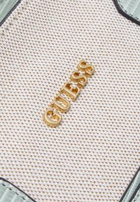 Tissu blanc texturé avec de petites perforations et lettres du logo "GUESS" en métal doré fixées, bordé de bords en cuir vert clair.
