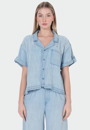 Junge Frau mit glattem blondem Haar, die ein hellblaues Jeans-Kurzarmhemd und passende hochgeschnittene Hose trägt.