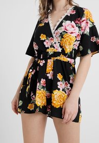 Mustat kukkakuosiset haalarit, joissa on värikkäitä keltaisia ja vaaleanpunaisia ruusuja, syvä V-pääntie, hihattomat hihat, kapea vyötärö ja pitsiyksityiskohtia.