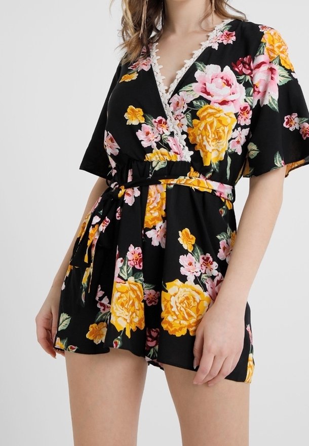 Mustat kukkakuosiset haalarit, joissa on värikkäitä keltaisia ja vaaleanpunaisia ruusuja, syvä V-pääntie, hihattomat hihat, kapea vyötärö ja pitsiyksityiskohtia.