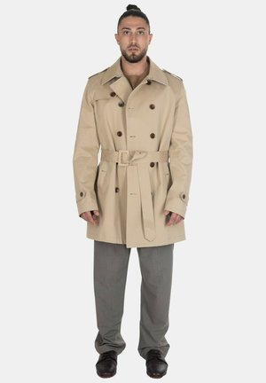 Uomo in piedi dritto che indossa un trench beige doppiopetto con cintura, pantaloni grigio e scarpe scure su uno sfondo uniforme.