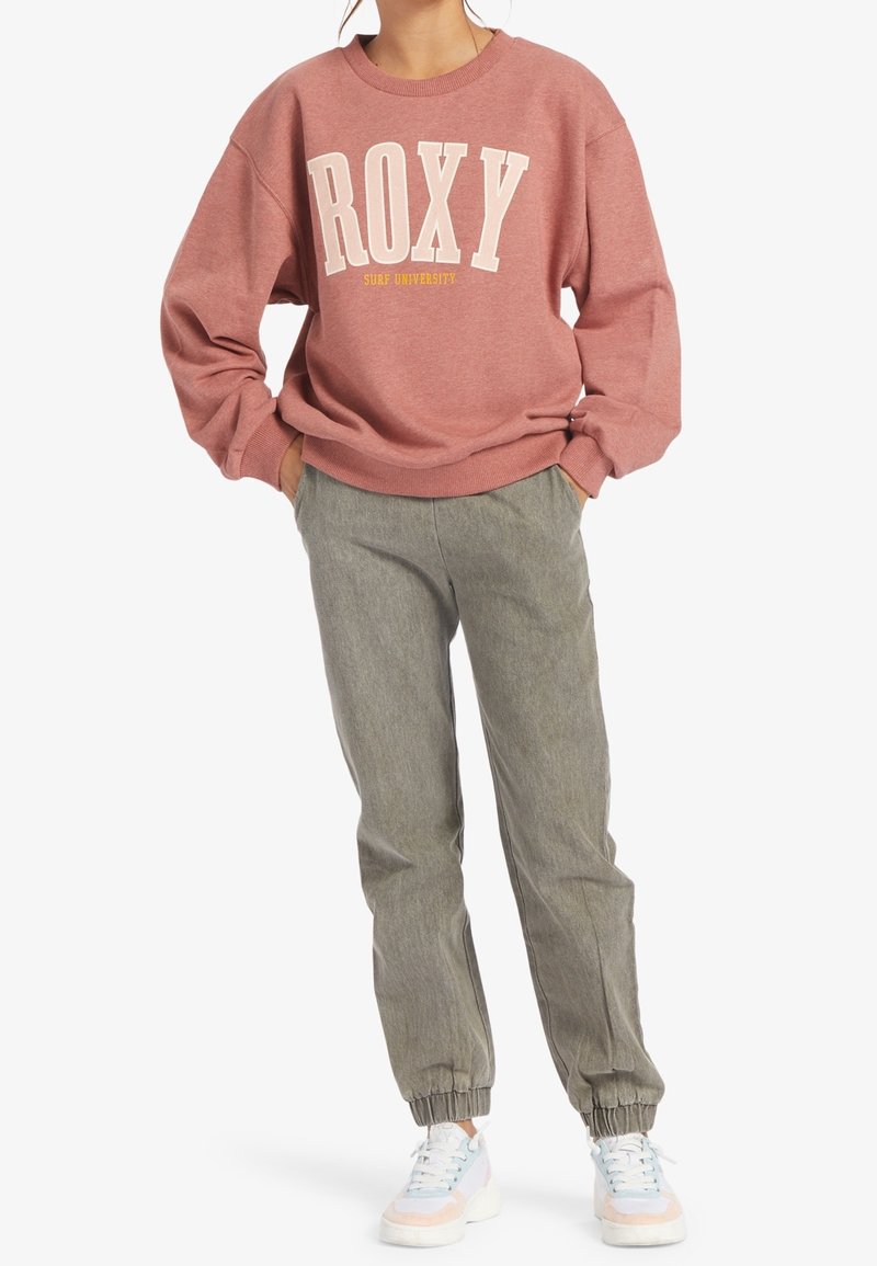 Femme portant un sweat-shirt rose poudré "ROXY Surf University", un pantalon de jogging gris et des baskets blanches avec des accents pastel. Mains dans les poches.