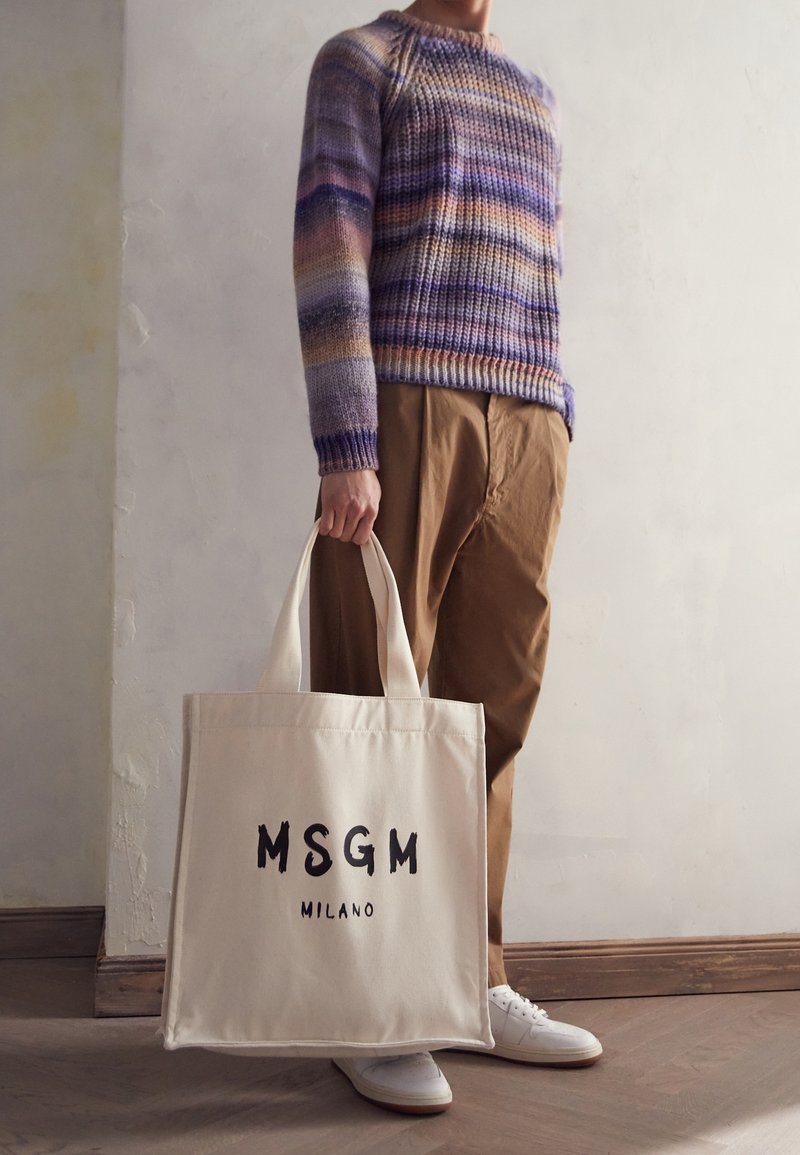Personne portant un pull rayé multicolore et un pantalon marron, tenant un sac cabas beige avec le texte "MSGM Milano", debout à l'intérieur.