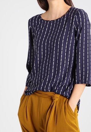 Navy blue top with white vertical dashed stripes and three-quarter sleeves paired with mustard yellow high-waist pants tied at the waist.