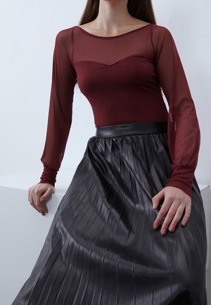 Femme portant un haut transparent bordeaux à manches longues et une jupe plissée noire taille haute, appuyée sur une surface blanche.