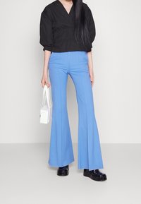 Svart kort topp med puffärmar, tillsammans med ljusblå utsvängda byxor. Modellen håller en liten vit handväska och bär svarta platåskor.