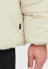 Puffjakke i cremefarve med en blank finish. Har en stramt samlet manchet og et sort label på sømmen. Blødt, tekstureret stof.