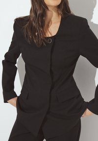 Zwarte op maat gemaakte blazer met een gestructureerd ontwerp, ronde gouden accentknop en twee voorzakken. Textuurstof en lange mouwen.