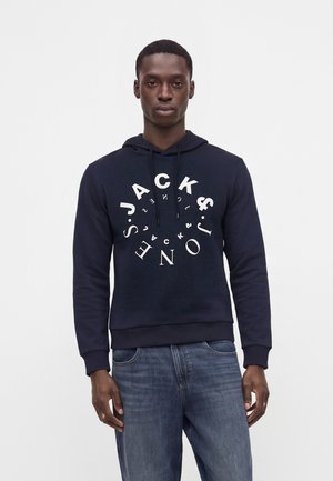 Młody mężczyzna w granatowej bluzie z kapturem z białym okrągłym napisem Jack & Jones oraz niebieskich dżinsach, stojący na jednolitym jasnym tle.
