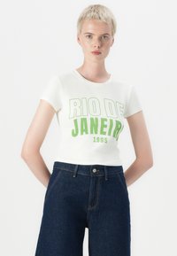 Hvit kortermet t-skjorte med fet grønn tekst "RIO DE JANEIRO 1995" på fronten, kombinert med mørkeblå høyhåndsjeans.