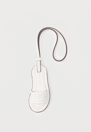 Biały skórzany brelok do kluczy w kształcie sandała z paskiem wokół kostki oraz wytłoczonym logo "Michael Kors" na przednim pasku, na jednolitym tle.
