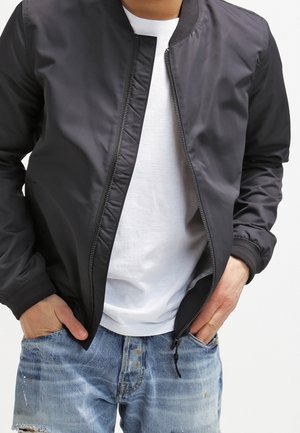 Person wearing a black zip-up bomber jacket over a white t-shirt and distressed blue jeans with hands in pockets.