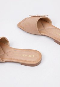 Sandalias deslizantes beige, fabricadas en un material similar al cuero suave, con una suela plana. Cuentan con una correa ancha y un detalle de acento dorado en la parte superior.