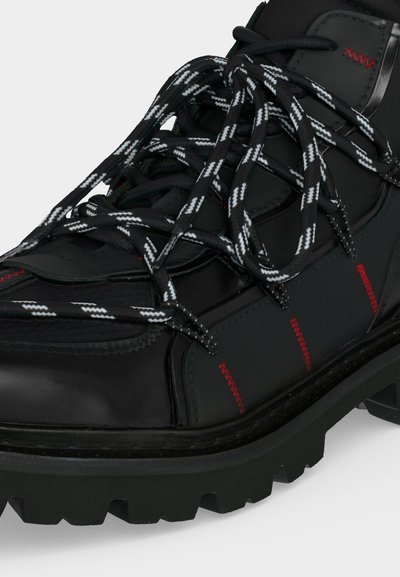 Gros plan d’une botte noire robuste avec des lacets épais blancs et noirs et des détails de couture rouge sur la tige et la semelle.