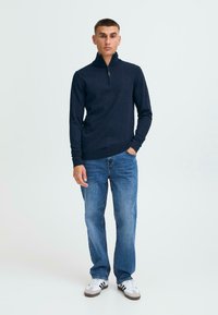 Pulover cu guler înalt de culoare navy, realizat dintr-un material tricotat, asortat cu blugi albaștri și adidași albi cu dungi negre. Design simplu și casual.