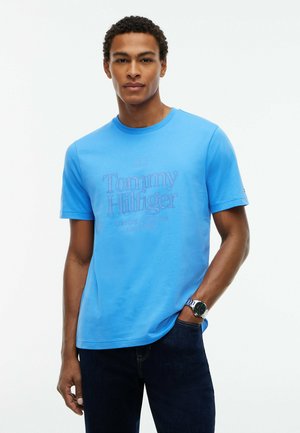 Man draagt heldere blauwe Tommy Hilfiger T-shirt en donkere spijkerbroek met een horloge aan de linkerpols, staande tegen een witte achtergrond.
