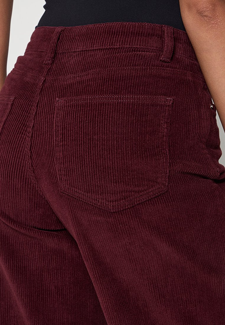 Bordeaux corduroy broek met verticale ribbels, voorzien van een achterzak en een standaard tailleband. Het materiaal heeft een zachte textuur.