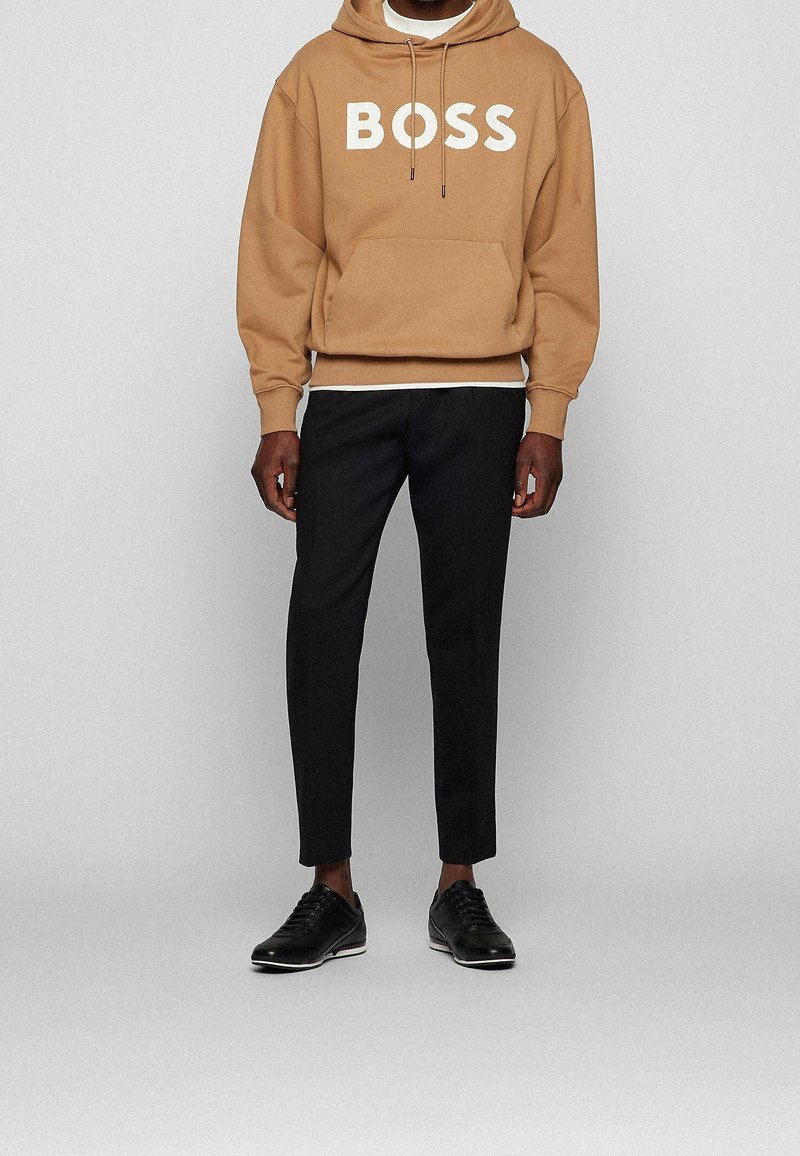 Brun huvtröja med vit "BOSS" text, i kombination med svarta smalrandiga byxor och svarta snörskor, som har en jämn yta.