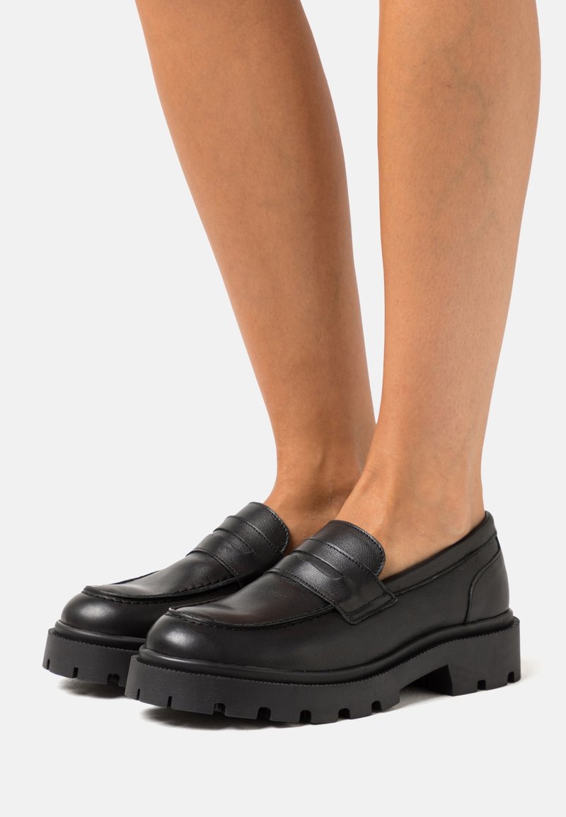 Personne portant des mocassins en cuir noir sans lacets avec des semelles épaisses en caoutchouc, pieds nus, debout devant un fond clair uni.