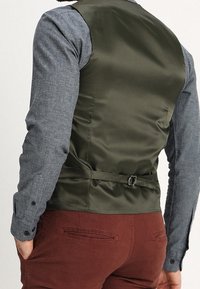 Gilet di raso verde oliva con retro regolabile, indossato sopra una camicia grigia a bottoni, abbinato a pantaloni marroni. Texture liscia, vestibilità sartoriale.
