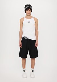 Hvit ärmeless tank topp med en svart logo, parret med svarte shorts med en Versace-midje, og hvite sneakers med svarte detaljer.