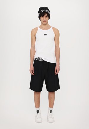 White sleeveless tank top with a black logo, paired with black shorts featuring a Versace waistband, and white sneakers with black accents.