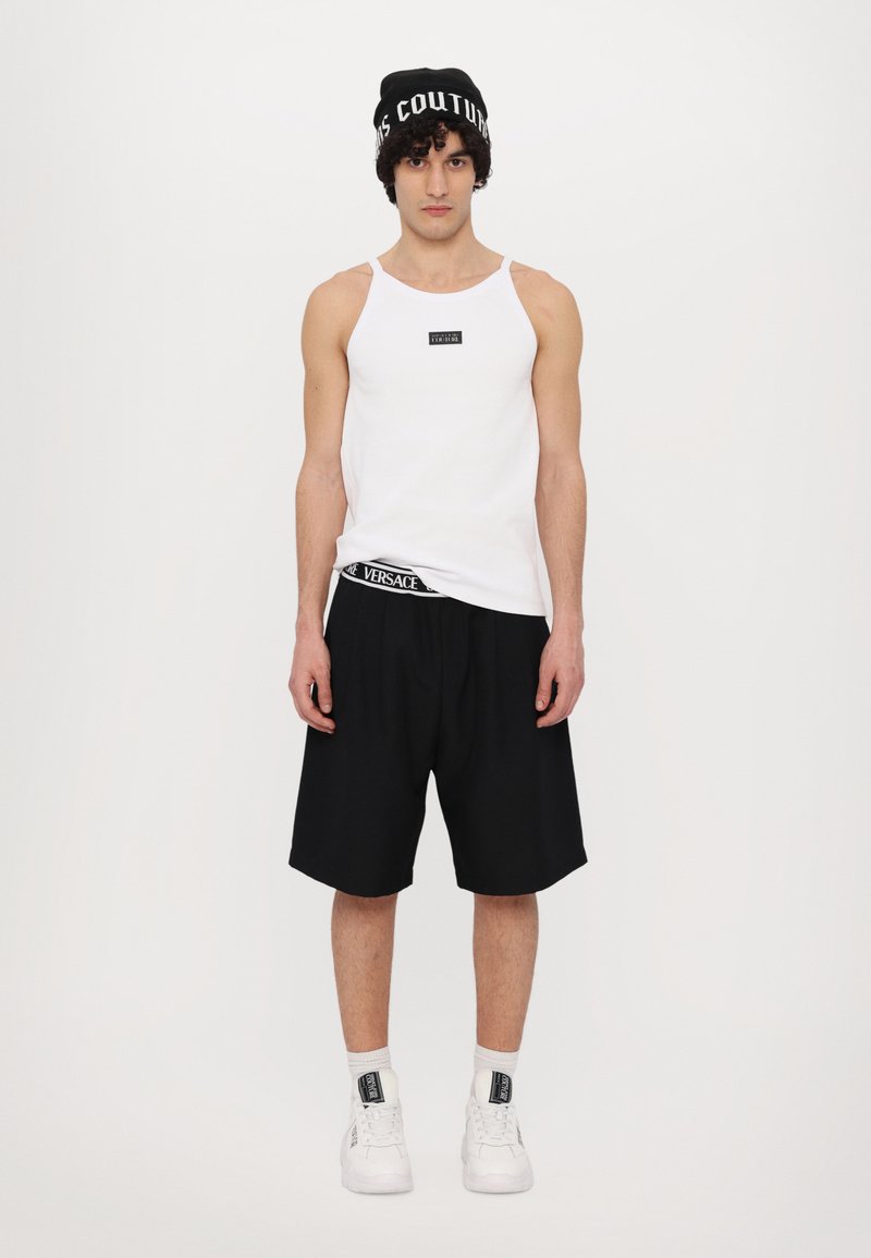 Hvit ärmeless tank topp med en svart logo, parret med svarte shorts med en Versace-midje, og hvite sneakers med svarte detaljer.