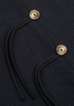 Zbliżenie czarnego teksturowanego materiału z dwoma beżowymi, okrągłymi guzikami oraz przeszywanymi, zakrzywionymi ozdobnymi pętlami.